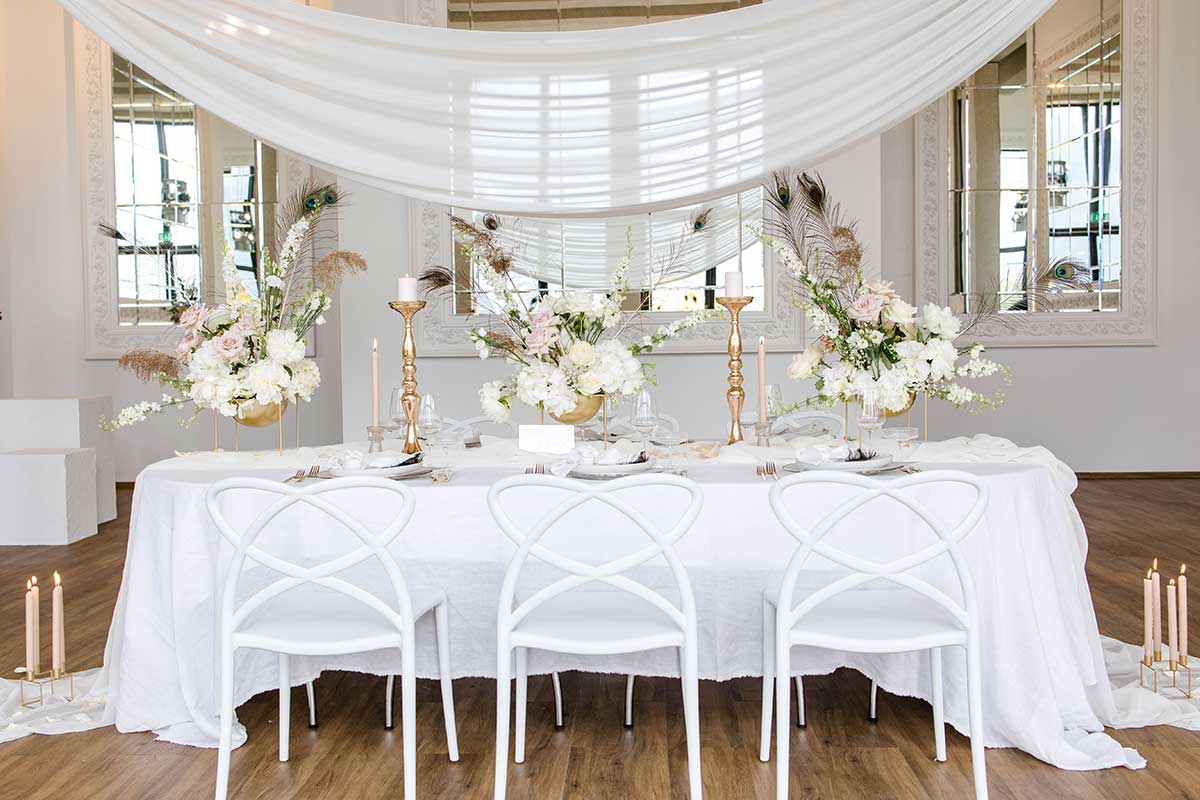Gedeckter Hochzeitstisch mit drappiertem Baldachin und weißen verschnörkelten Stühlen